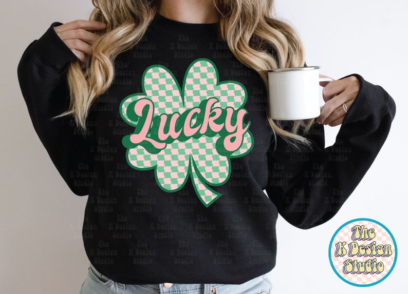May include: Black sweatshirt featuring a large, retro-style shamrock graphic with a checkered pattern in green and pink. The word "Lucky" is written across the shamrock in a cursive font. A white mug is held in the image.