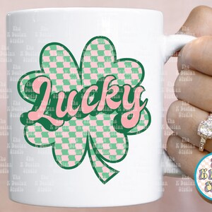 May include: White ceramic mug featuring a green and pink checkered shamrock design with the word "Lucky" in pink script. The mug is held by a hand with a ring. The K Design Studio logo is visible.