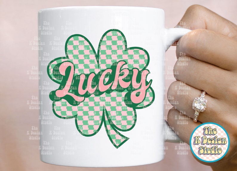 May include: White ceramic mug featuring a green and pink checkered shamrock design with the word "Lucky" in pink script. The mug is held by a hand with a ring. The K Design Studio logo is visible.