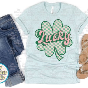 May include: Light blue t-shirt with a green and pink checkered shamrock design and the word "Lucky" in pink. The shirt is paired with blue jeans and silver sandals. The K Design Studio logo is visible.