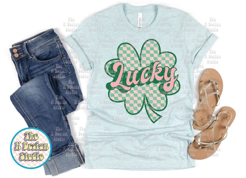May include: Light blue t-shirt with a green and pink checkered shamrock design and the word "Lucky" in pink. The shirt is paired with blue jeans and silver sandals. The K Design Studio logo is visible.