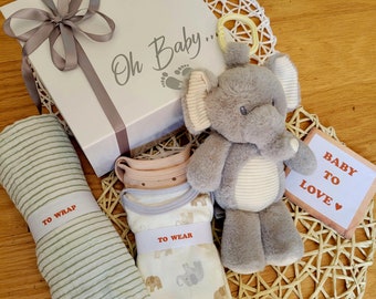 Può includere: Un cesto regalo per neonati con una scatola bianca che dice "Oh Baby..." con un peluche di elefante grigio, una coperta a righe e un body bianco con stampa di elefante. Il cesto include anche un cartello di legno che dice "Baby to Love".