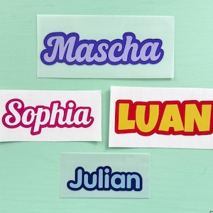 Op de afbeelding: Vier rechthoekige gepersonaliseerde naamstickers met de namen Mascha in paars, Sophia in roze, LUAN in geel met een rode rand en Julian in blauw. De tekst Personalisierte Namenssticker staat bovenaan en Persoenlich. Kreativ. Selbstgemacht. onderaan.