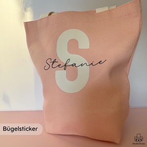 Könnte beinhalten: Eine hellrosa Canvas-Tragetasche mit cremefarbenen Griffen. Die Tasche zeigt einen großen weißen Buchstaben "S" und den Namen "Stefanie" in Schreibschrift. Ein schlichtes, stilvolles Accessoire.
