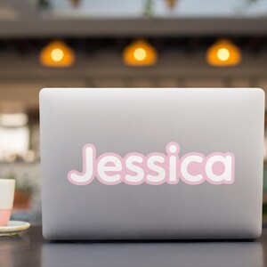 Op de afbeelding: Een zilverkleurige laptop met een aangepaste vinylsticker op de deksel. De sticker toont de naam Jessica in een afgerond, vet wit lettertype met een zachtroze omlijning, wat zorgt voor een strakke en goed leesbare uitstraling op het oppervlak.