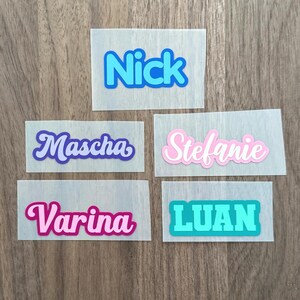 Op de afbeelding: Vijf rechthoekige zelfklevende labels met namen in verschillende lettertypes en kleuren. De labels tonen de namen Nick in blauw, Mascha in paars, Stefanie in roze, Varina in magenta en LUAN in taling op een doorschijnende achtergrond.