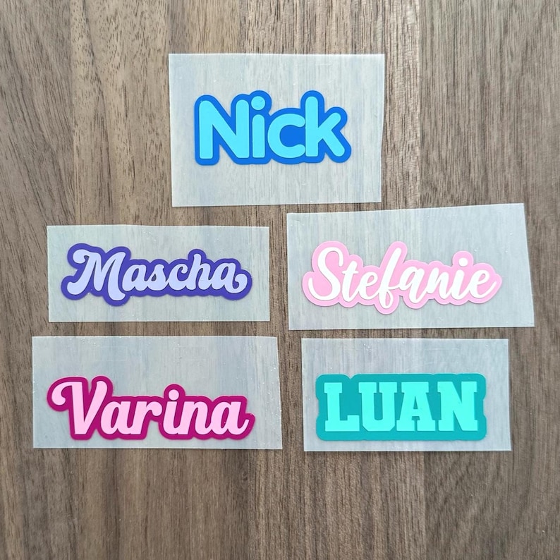 Op de afbeelding: Vijf rechthoekige zelfklevende labels met namen in verschillende lettertypes en kleuren. De labels tonen de namen Nick in blauw, Mascha in paars, Stefanie in roze, Varina in magenta en LUAN in taling op een doorschijnende achtergrond.