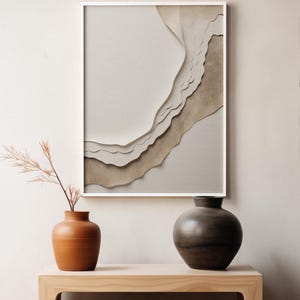 Könnte beinhalten: Zwei Keramikvasen, eine braune und eine dunkelgraue, stehen auf einem hellen Holztisch vor einem gerahmten abstrakten Kunstdruck mit weißem Hintergrund und übereinanderliegenden braunen Papierformen.
