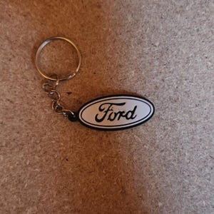May include: A keychain featuring the Ford logo. The logo is an oval shape with a white background and black border, with the word "Ford" in a cursive font. The keychain has a silver metal ring and chain.