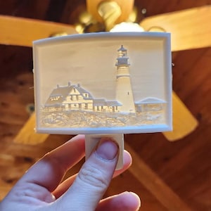 May include: A white night light featuring a detailed relief of a lighthouse and adjacent buildings. The design is rectangular with a plug-in base. The lighthouse is tall and cylindrical, with a light source at the top. The buildings are low and rectangular.