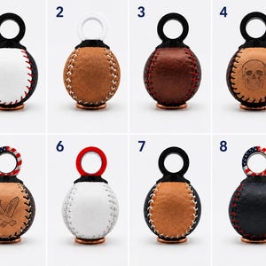 Könnte beinhalten: Acht kugelförmige Schlüsselanhänger aus Leder mit runden Schlaufen, nummeriert von eins bis acht. Die Designs umfassen Baseball-Nähte, eine Totenkopf-Gravur, eine Adler-Gravur und US-Flaggenmuster an den Schlaufen in verschiedenen Lederfarben.