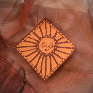 Può includere: Un sottobicchiere quadrato arancione con un disegno di sole. Il sole ha un volto sorridente e raggi, delineati in marrone. Il sottobicchiere è posto su un tessuto strutturato, arancione-rossastro.