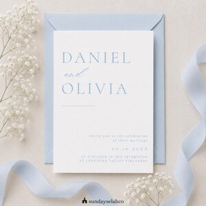 May include: A white wedding invitation card with light blue text featuring the names DANIEL and OLIVIA, placed inside a light blue envelope. The card includes event details, date 09.10.20XX, and location Canungra Valley Vineyards.