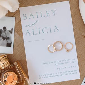 K&ouml;nnte beinhalten: Eine wei&szlig;e Hochzeitseinladungskarte mit hellblauem Text, der BAILEY and ALICIA, invite you to the celebration of their marriage, 09.10.2025, at 3 o'clock in the afternoon at Canungra Valley Vineyards lautet, mit drei goldenen Ringen.