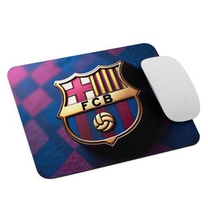 May include: A rectangular mousepad featuring the FC Barcelona crest in gold, red, and blue. The crest is centered on a background of blue and purple diamond pattern. A white computer mouse is positioned to the right of the mousepad.