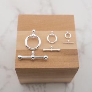 May include: A set of silver-toned metal jewellery findings, including a ring, a bar, and a toggle clasp. The findings are arranged on a wooden surface.