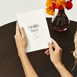 Può includere: Un libretto bianco con il testo "Fertility Planner" sulla copertina. Il libretto è tenuto da una persona con le mani visibili.