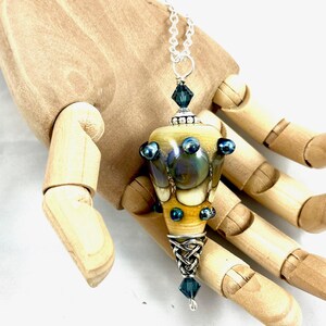 May include: A silver chain necklace with a large, yellow, blue, and iridescent glass pendant. The pendant is shaped like a cone and has a silver Celtic knot design at the bottom. The pendant is accented with small blue glass beads.