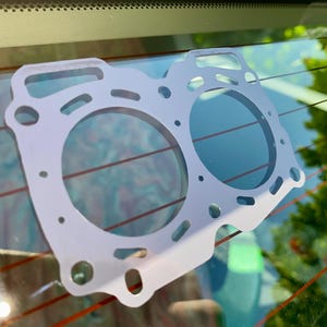 May include: A light gray head gasket with two large circular openings and several smaller holes. The gasket has an irregular shape with rounded edges and is designed for automotive use.