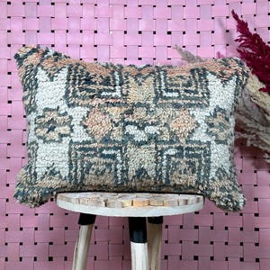 May include: A rectangular throw pillow with a geometric pattern in shades of beige, brown, and gray. The pillow is sitting on a wooden stool against a pink woven background.