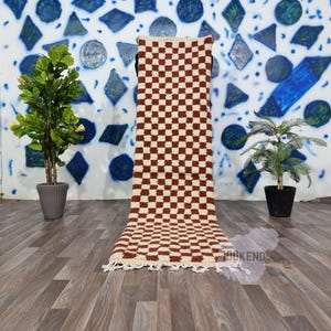 May include: A brown and white checkered rug with fringe on the edges. The rug is laid out on a wooden floor in front of a wall with blue and white geometric shapes.