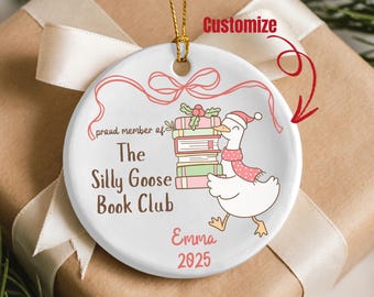 Pode incluir: Enfeite de Natal redondo em cerâmica branca com fio dourado. Apresenta um ganso de desenho animado com gorro e cachecol de Papai Noel, carregando livros. O texto inclui "The Silly Goose Book Club", "Emma 2025" e "Personalizar".