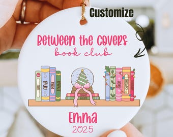 Pode incluir: Enfeite redondo de cerâmica branca com o texto rosa "Between the covers book club". O enfeite apresenta um design de estante com livros coloridos, um globo de neve e o nome "Emma" com o ano de 2025.