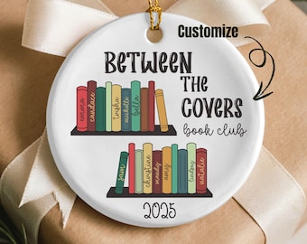 Può includere: Un ornamento rotondo in ceramica bianca con un anello dorato per appenderlo. L'ornamento presenta il testo "Between The Covers book club" e l'anno "2025". Mostra due file di libri colorati con nomi sui dorsi.