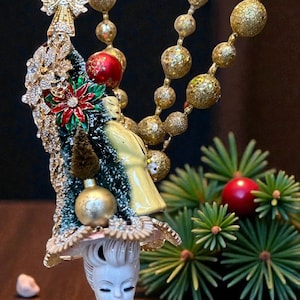 May include: A vintage Christmas ornament featuring a porcelain lady figurine with a hat adorned with a miniature Christmas tree, angel, and red poinsettia. Gold beaded garland and a sprig of evergreen complete the festive scene.