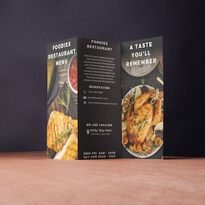 May include: A tri-fold menu for Foodies Restaurant. The front panel features the text "A Taste You'll Remember" and a photo of a roasted chicken with garlic and herbs. The back panel has the restaurant's name, address, phone number, and hours of operation.