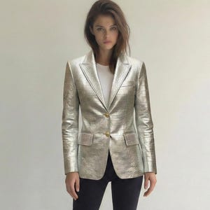 May include: A metallic silver blazer with a textured finish, featuring a notched lapel and two front pockets. The blazer has a single gold button closure. The person is wearing a white t-shirt, black pants, and brown ankle boots.