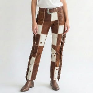 Könnte beinhalten: Braune und weiße Patchwork-Lederhose mit Fransen an den Seiten. Die Hose hat einen braunen Ledergürtel mit silberner Schnalle. Das Design ist eine Mischung aus braunen und weißen Quadraten.