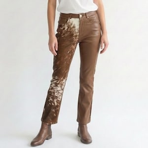 Könnte beinhalten: Braune Lederhose mit einem Kuhfell-Einsatz am linken Bein. Die Hose hat ein klassisches Fünf-Taschen-Design und eine gerade Beinpassform. Das Leder ist in einem satten, warmen Braunton gehalten, und der Kuhfell-Einsatz sorgt für Textur.