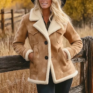 May include: A tan suede jacket with a cream shearling collar and trim. The jacket features two chest pockets, two lower pockets, and black buttons. The model is wearing a black hat and dark trousers. A classic outerwear item.