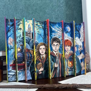 Könnte beinhalten: Ein Buchset mit Buchschnittmalerei, das eine durchgehende Illustration von Harry-Potter-Charakteren, einem Dampfzug, einem Schloss und einer Eule zeigt. Das Kunstwerk zeigt Hermine, Harry und Ron in Schuluniformen vor einem blauen Wald.