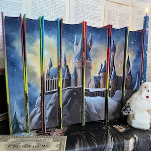 Può includere: Una collezione di libri con copertina rigida con bordi e dorsi dipinti su misura. I dorsi sono dipinti con un cielo notturno e una scena di castello. Sono visibili una statuina di gufo bianco e una candela blu.