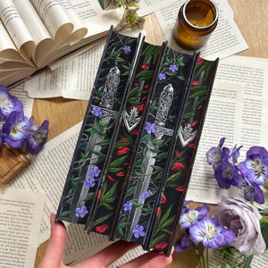 Könnte beinhalten: Vier Bücher mit schwarzen Buchrücken, verziert mit einem silbernen Schwert, roten Blütenblättern, grünen Blättern und lila Blumen. Die Bücher liegen auf einem Tisch mit offenen Büchern und Blumen.