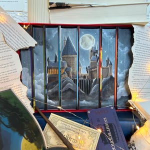 Könnte beinhalten: Ein Set aus sieben Büchern in einem roten Rahmen, die auf ihren Buchrücken eine durchgehende Illustration einer nächtlichen Burg unter einem Vollmond zeigen. Die Szene umfasst felsiges Gelände, Wolken, Sterne sowie ein Ticket und einen Zauberstab.