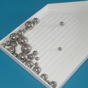 Peut inclure: Un plateau en plastique blanc rempli de billes métalliques argentées brillantes de différentes tailles. Les billes sont disposées en diagonale, certaines étant dispersées sur le plateau. Le fond est de couleur turquoise unie.