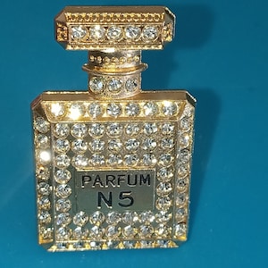 Pode incluir: Broche dourado em forma de frasco de perfume, incrustado com strass transparentes. O broche tem um corpo retangular com uma parte superior quadrada e um pescoço pequeno e redondo. O texto "PARFUM N5" é exibido na frente.