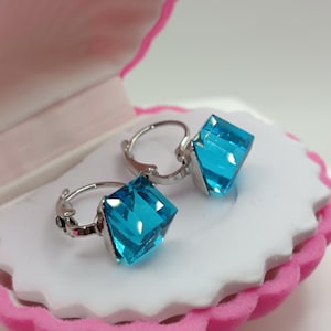 Peut inclure: Boucles d'oreilles créoles en argent avec des pierres précieuses carrées bleu clair. Les boucles d'oreilles sont présentées dans un écrin rose et blanc en forme de coquillage.