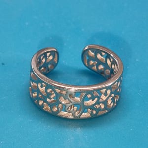 Könnte beinhalten: Silberfarbener offener Ring mit einem aufwendigen, durchbrochenen floralen Design. Der Ring ist vor einem einfarbigen blauen Hintergrund platziert, der das detaillierte Muster hervorhebt. Dieser verstellbare Ring ist ein Schmuckstück.