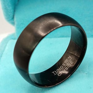 Può includere: Un anello nero in carburo di tungsteno con finitura spazzolata. L'anello ha un design liscio e arrotondato e le parole "TUNGSTEN CARBIDE" sono incise all'interno. L'anello è esposto su una superficie azzurra.