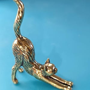 Op de afbeelding: Een bronzen kattenbeeldje in een rekpositie. De kat staat op alle vier de poten, met de voorpoten naar voren uitgestrekt en de staart omhoog gebogen. Het beeldje heeft een gedetailleerde vachttextuur en staat tegen een blauwe achtergrond.