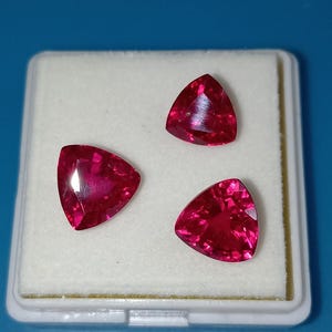 Op de afbeelding: Drie driehoekige, gefacetteerde, dieprode edelstenen worden getoond in een witte plastic doos tegen een blauwe achtergrond. De edelstenen hebben een hoge glans en reflecteren licht. De vorm is een driehoek met afgeronde hoeken. Dit kunnen robijnen zijn.