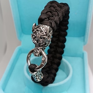 Può includere: Bracciale nero intrecciato con chiusura a testa di pantera argentata e dettagli decorativi. Il bracciale è presentato in un cofanetto azzurro, che mette in risalto i dettagli intricati.