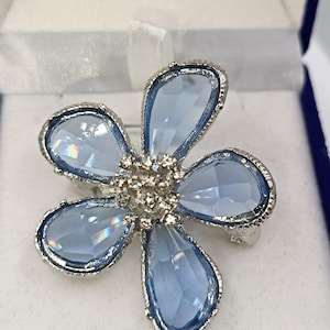 Pode incluir: Um broche em forma de flor com cinco pétalas facetadas azul claro. As pétalas são contornadas em prata, e o centro do broche é um aglomerado de metal prateado. O broche é exibido em uma caixa branca e azul.