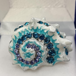 Könnte beinhalten: Eine dekorative Schmuckschatulle in Form einer Muschelschale. Die weiße Schale ist mit türkisfarbenem Emaille akzentuiert und mit blauen und lila Edelsteinen verziert. Die Box wird in einer weiß-blauen Box präsentiert, was auf ein Geschenk hindeutet.