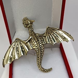 Può includere: Spilla a forma di drago color oro con ali spiegate, presentata in un portagioie rosso e bianco. Il drago presenta squame e motivi alari dettagliati ed è realizzato in metallo lucido.