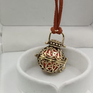 Peut inclure: Un collier pendentif filigrané de couleur or avec un intérieur rouge. Le pendentif est rond et suspendu à un cordon marron. Le collier est présenté dans un écrin blanc.